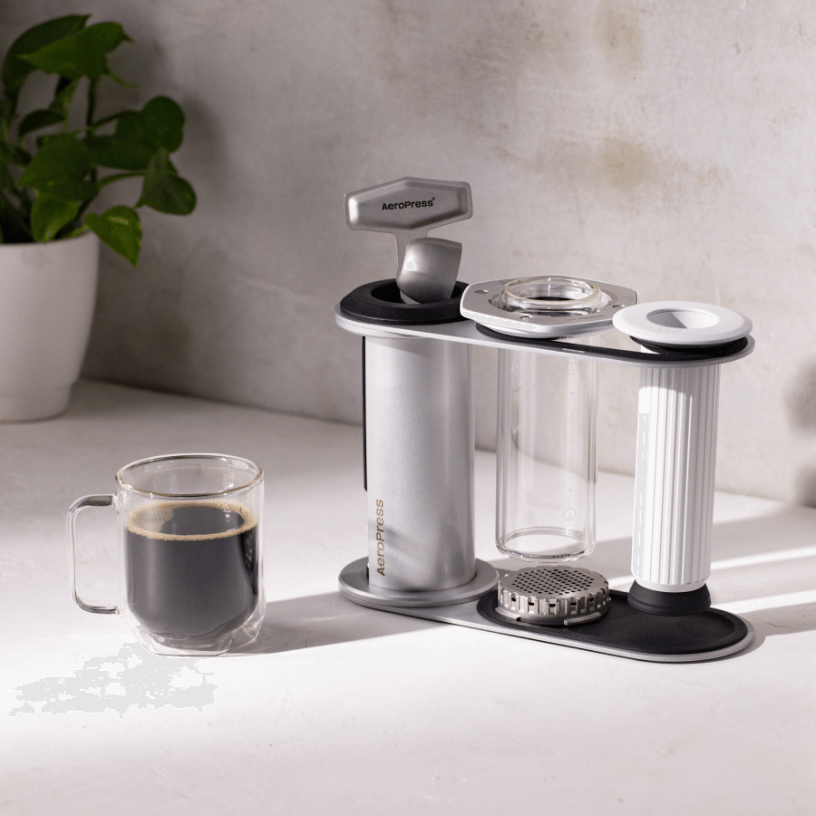 White AeroPress Premium in the stand on a white surface with a cup of coffee and plant in the background. #color_white
