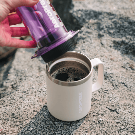 Person pouring coffee from a purple AeroPress into a white mug on a concrete surface. #color_purple