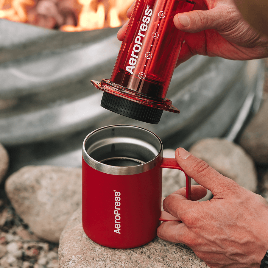 Person using a red Aeropress coffee maker over a red mug outdoors. #color_red