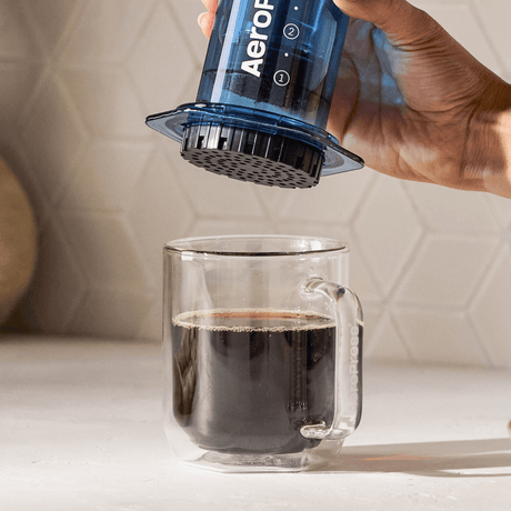 A person using an Aeropress coffee maker to pour coffee into a clear glass mug. #color_blue