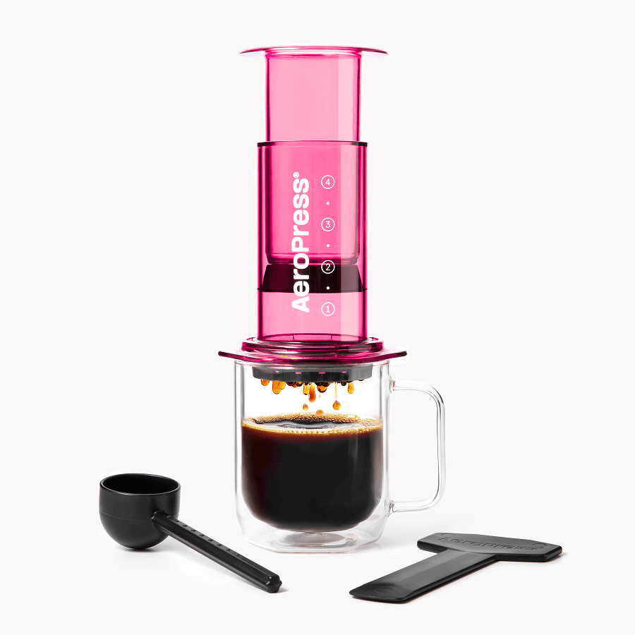 Pink AeroPress coffee maker with a glass mug and black scoop on a white background #color_pink