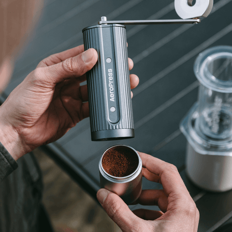 Person using an AeroPress manual coffee grinder with a focus on the coffee grounds.
