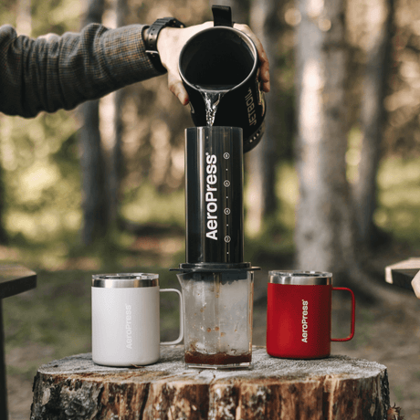 Person pouring coffee from an Aeropress into a mug outdoors.