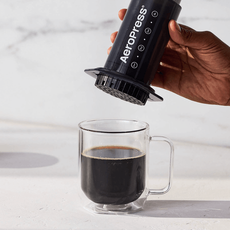 A person using an Aeropress to pour coffee into a clear glass mug on a light background