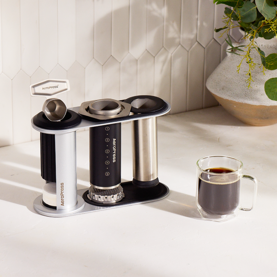 Aeropress coffee maker with three pods and a glass of coffee on a light surface. #color_black