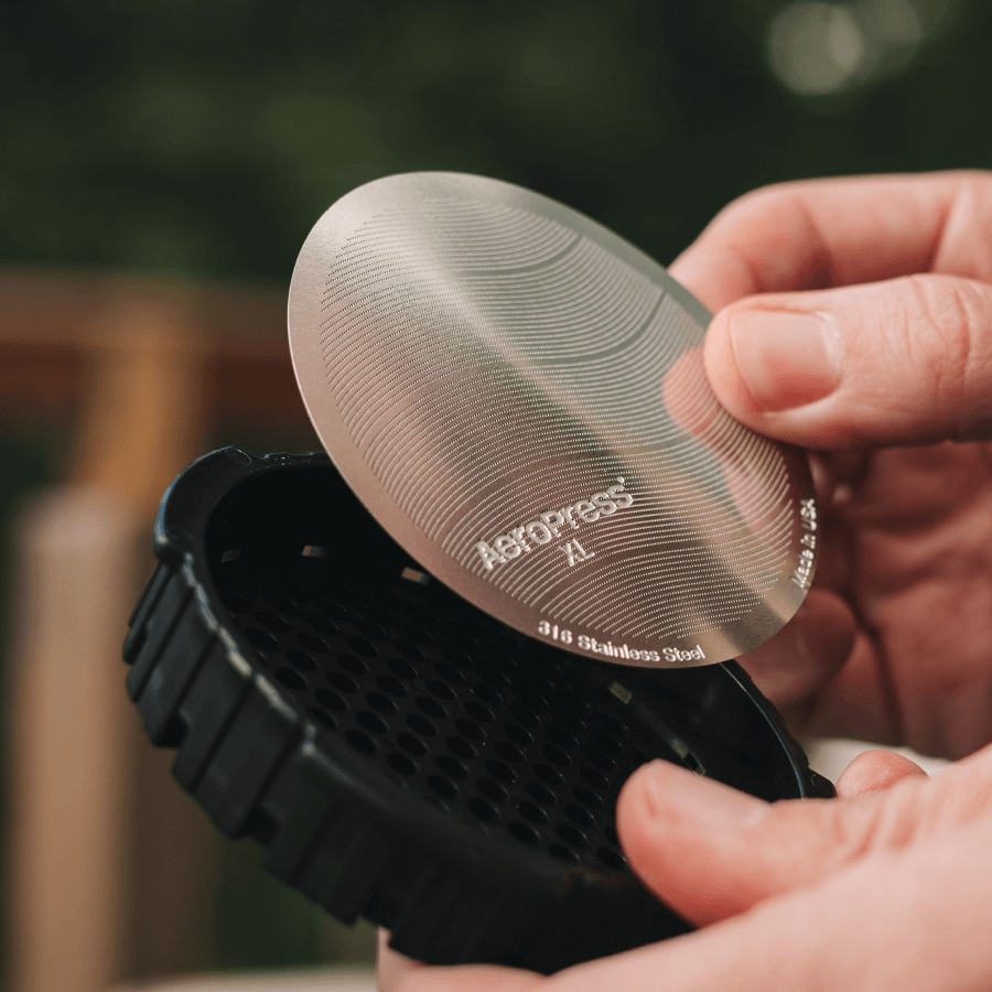 Person holding a black AeroPress coffee maker with a metal filter labeled 'AeroPress'.