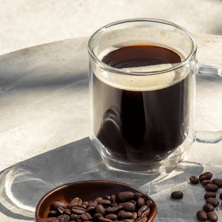 Clear glass mug filled with coffee, on a reflective surface with scattered coffee beans.