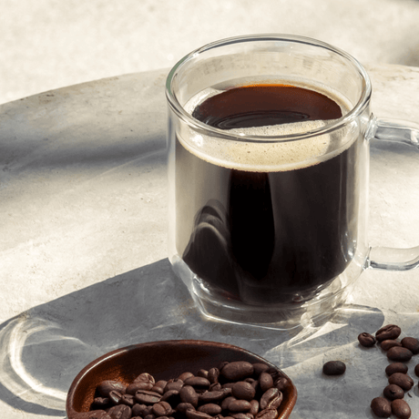 Clear glass mug filled with coffee, on a reflective surface with scattered coffee beans.
