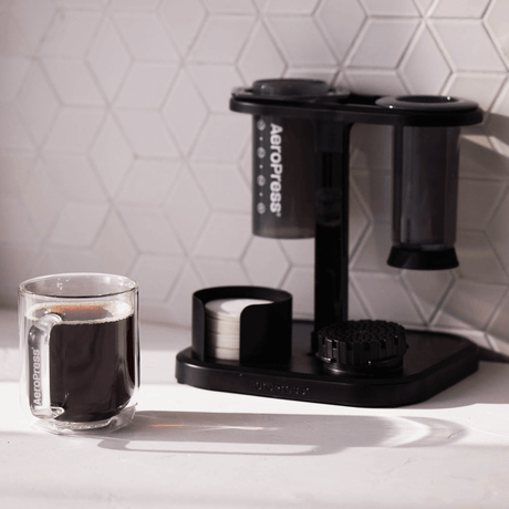 Aeropress coffee maker with a glass of coffee on a white surface.