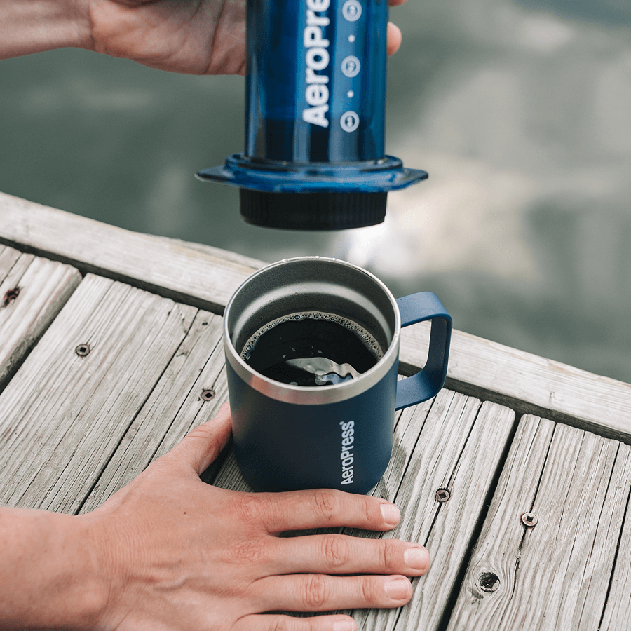 Person using an AeroPress to make coffee on a wooden deck by a body of water. #color_blue