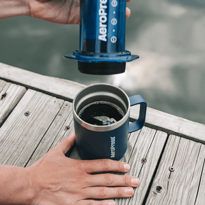 Person using an AeroPress to make coffee on a wooden deck by a body of water. #color_blue