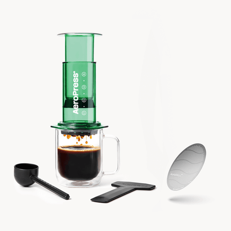 Green AeroPress coffee maker with a glass mug of coffee and accessories on a white background #color_green
