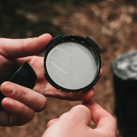 Person holding a coffee filter with a brand name against a blurred natural background #color_green