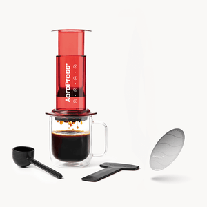 Red AeroPress coffee maker with a clear mug, black scoop, and filter on a white background #color_red