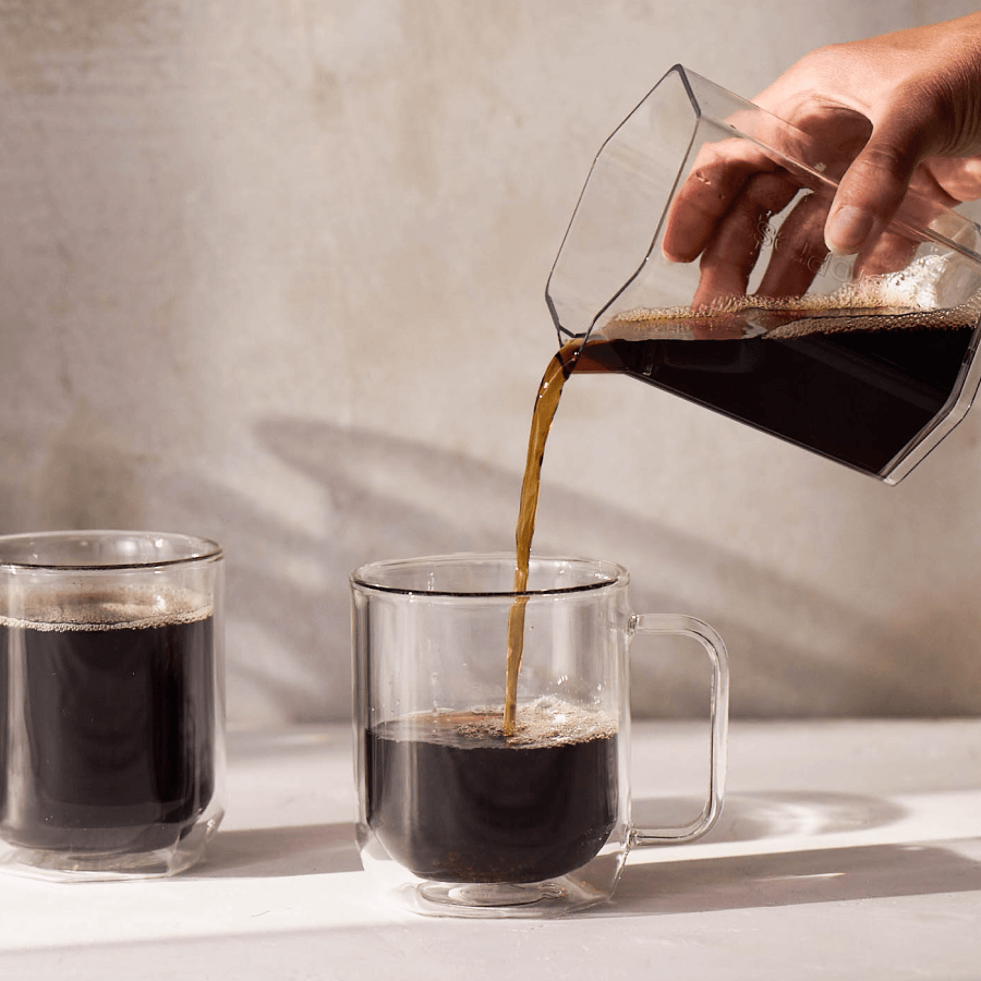 Person pouring coffee from a carafe into a clear mug on a light surface.