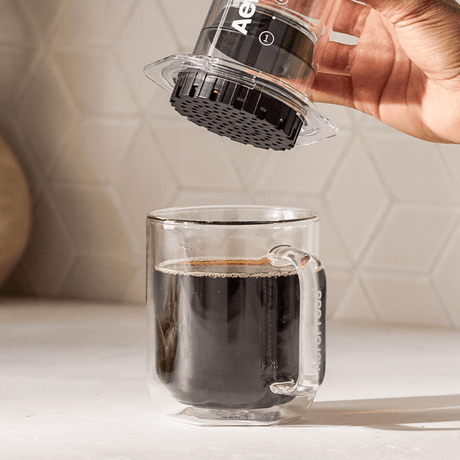 Hand pouring coffee from a clear glass container into a glass mug on a light surface.