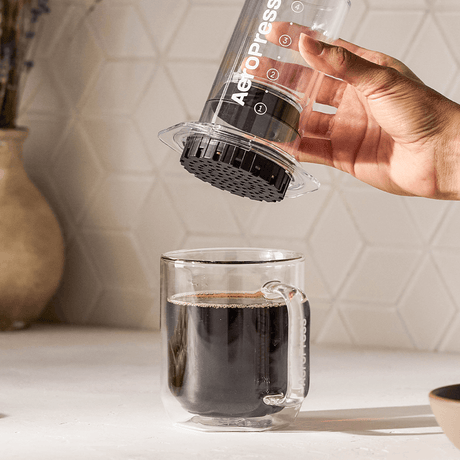 Hand using an Aeropress to make coffee in a clear glass mug on a kitchen counter. #color_clear