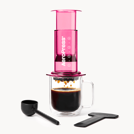 Pink AeroPress coffee maker with a glass mug and black scoop on a white background #color_pink