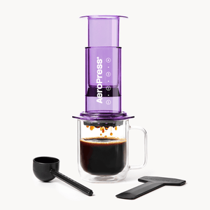 Purple AeroPress coffee maker with a glass mug of coffee and scoop on a white background #color_purple