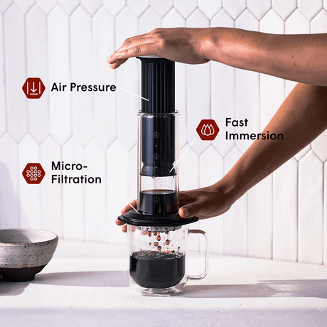 Person using an AeroPress Premium Black coffee maker with text labels for air pressure, fast immersion, and micro-filtration. #color_black