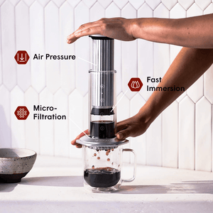 Person using an AeroPress Premium coffee maker with text labels on air pressure, fast immersion, and micro-filtration. #color_silver