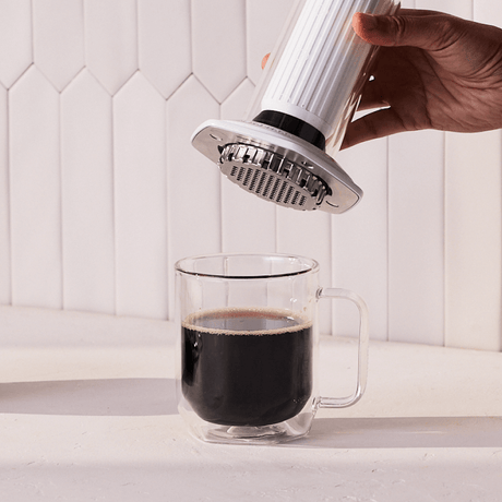 Hand using an AeroPress Premium coffee maker to pour coffee into a clear glass mug on a white countertop. #color_white