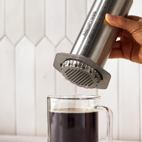 Hand using a silver AeroPress to press coffee into a glass mug with a white tiled wall background #color_silver
