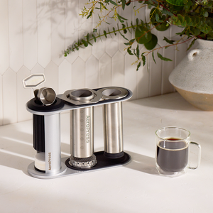 Aeropress coffee maker on a kitchen counter with a cup of coffee and a plant in the background. #color_silver