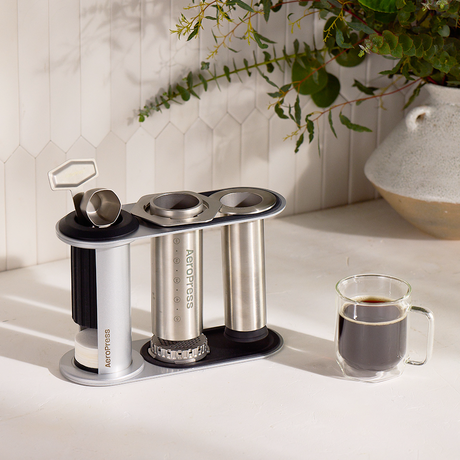 Aeropress coffee maker on a kitchen counter with a cup of coffee and a plant in the background. #color_silver