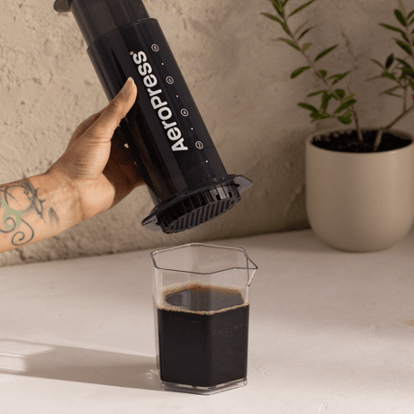 A person using an Aeropress to make coffee, with a glass of coffee on a table.